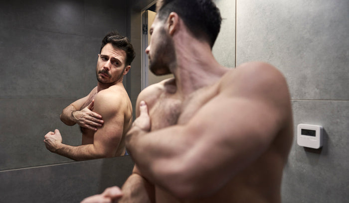 Muskulöser Mann betrachtet sich kritisch im Spiegel – Fitness und Körperbewusstsein im Badezimmer